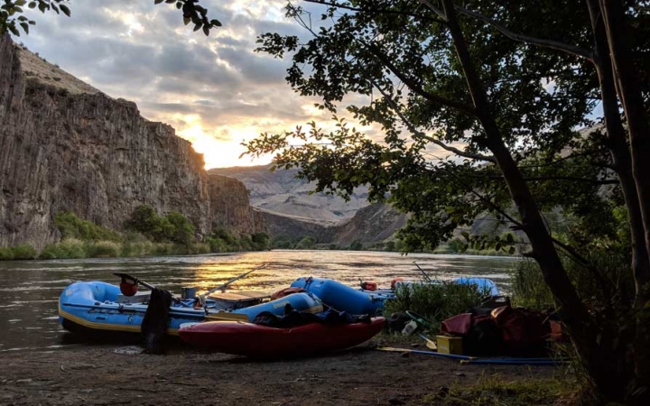 Rafts and kayaks rest on the shore of a calm river, In the background, the river is framed by high canyon walls.